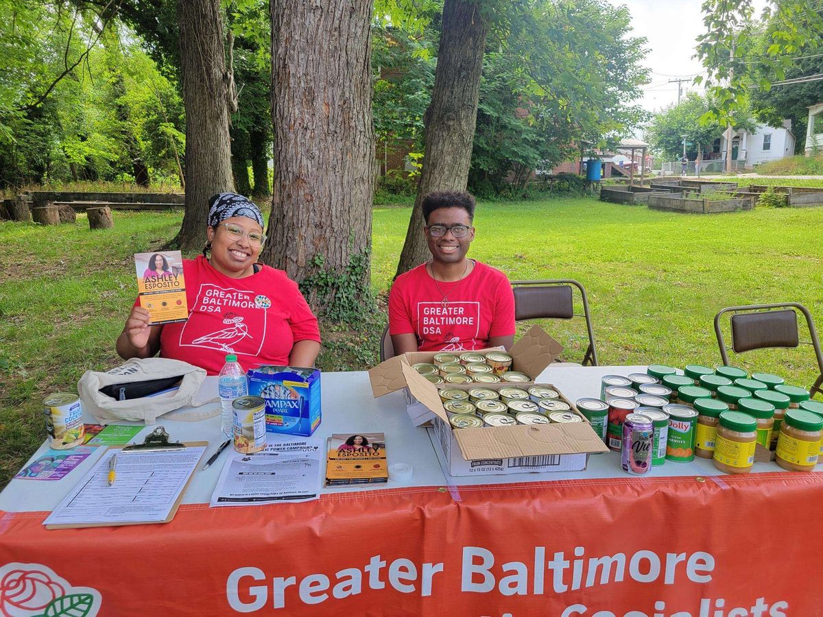Two members of color sitting at a table with GBDSA banner and several cans/jars of food and other resources available. Member on the left is holding a brochure for Ashley Esposito's re-election campaign for school board.