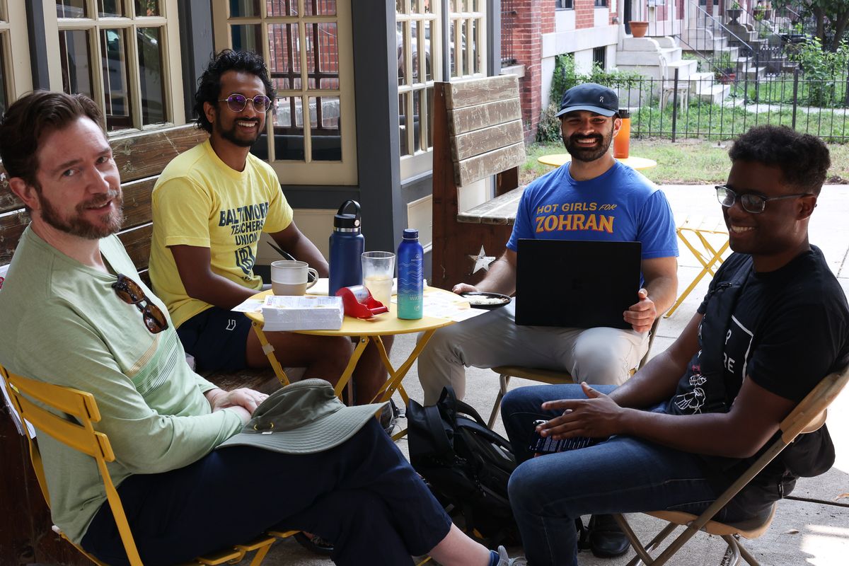 Four GBDSA members at a small cafe table outside. One is wearing a Baltimore Teachers Union shirt, and another is wearing a Hot Girls for Zohran shirt.