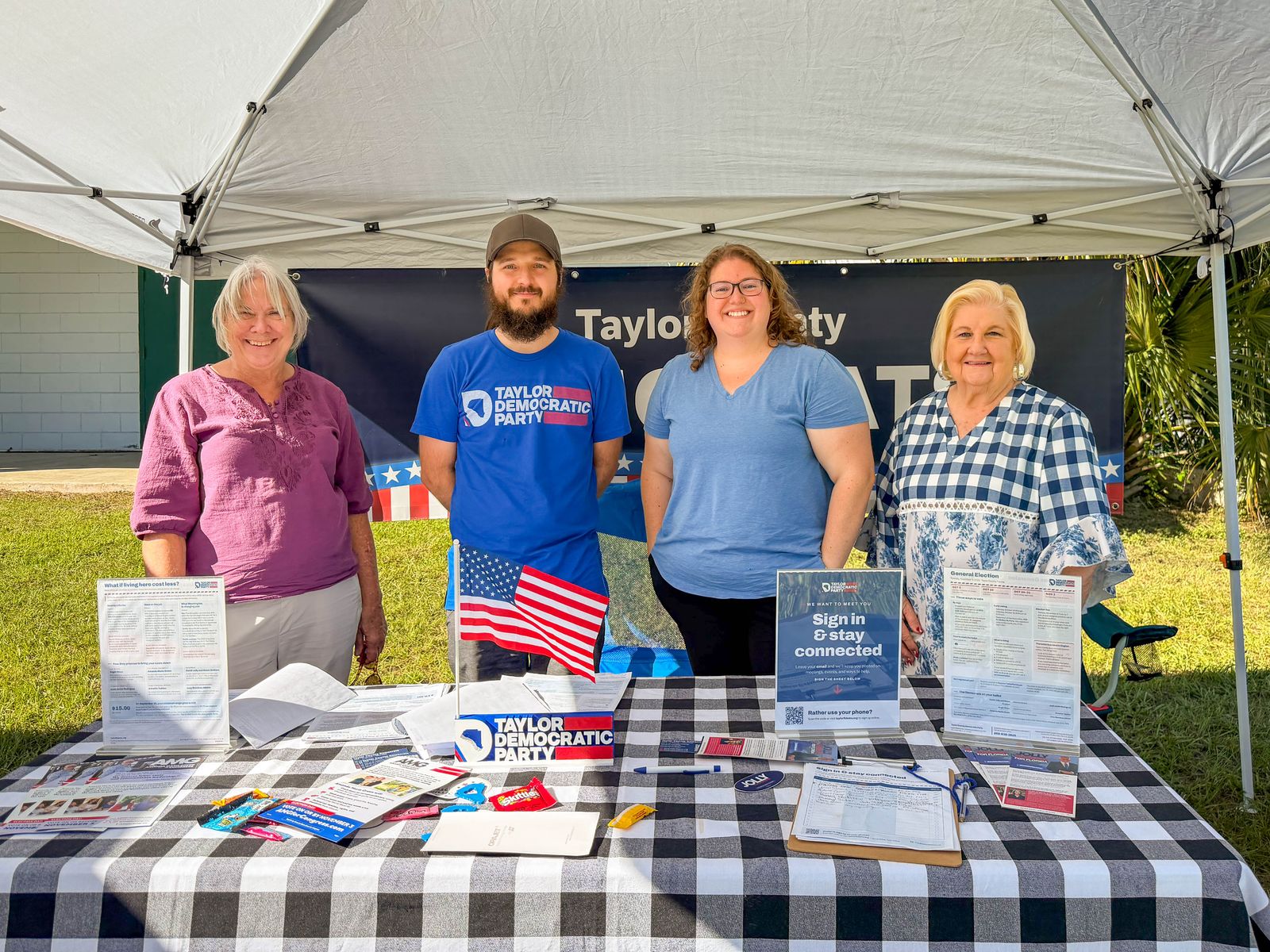 Four Taylor County Democrats volunteers smiling behind the party's information table under a white tent at Smokin' in the Pines