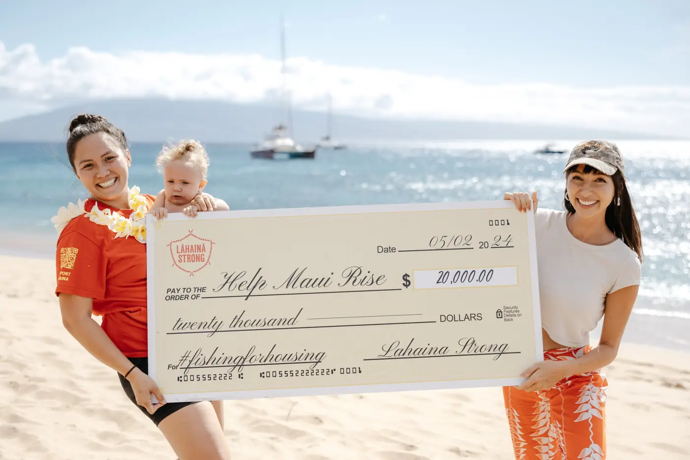 Two women on a Lāhainā beach holding a large donation check, smiling